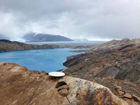 Blick über braune Hügel auf einen See, dahinter ein Gletscher