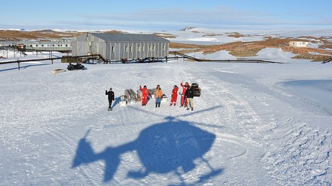 Eine Eisfläche, inmitten eine Gruppe Wissenschaftler. Darüber sieht man den Schatten eines Helikopters.