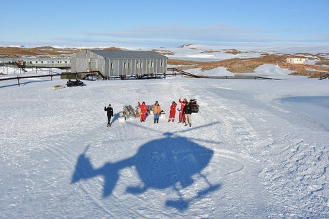 Eine Eisfläche, inmitten eine Gruppe Wissenschaftler. Darüber sieht man den Schatten eines Helikopters.