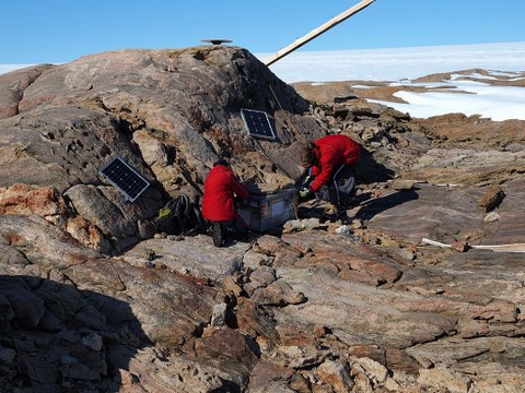 Ein steiniger Hügel, darauf hocken in roten Schutzanzügen zwei Wissenschaftler. 