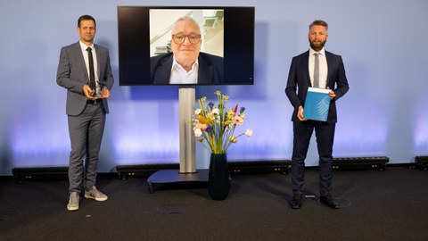 Zwei Herren mit Urkunden in der Hand, in der MItte ein älterer Herr auf einem Flachbildschirm. Davor eine Vase mit einem Blumenstrauß.