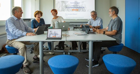 Um einen Tisch sitzen 5 ältere Personen vor Laptops.