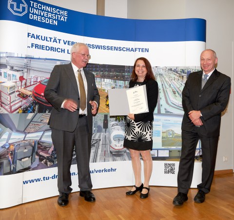 Zwei Männer und eine Frau in der Mitte stehen vor einer Fotowand der Fakultät Verkehrswissenschaften. Die Frau hält eine Urkunde in der Hand.