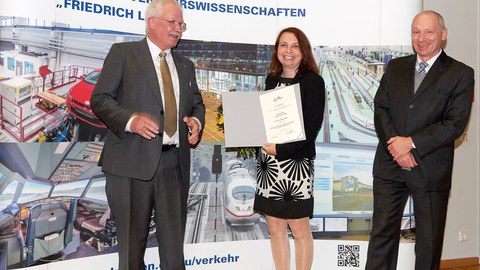 Zwei Männer und eine Frau in der Mitte stehen vor einer Fotowand der Fakultät Verkehrswissenschaften. Die Frau hält eine Urkunde in der Hand.