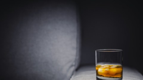 Foto von einem Glas Whiskey mit Eis, das auf einer grauen Sofalehne steht.