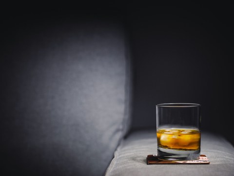 Foto von einem Glas Whiskey mit Eis, das auf einer grauen Sofalehne steht.