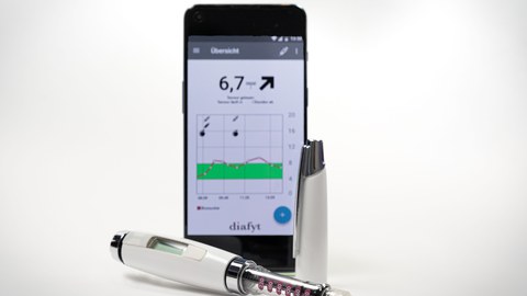 In der Mitte ein gerade stehendes Handy, auf dem Display die geöffnete diafyt-App. Davor liegt eine geöffnete Diabethes-Spritze (Pen).