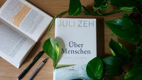 Auf einem Tisch liegt das Buch "Über Menschen" von Juli Zeh, links daneben ein aufgeschlagenes, orange gemarkertes Buch, rechts eine Rankpflanze.