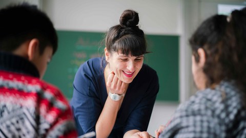 Eine Lehrerin stütze sich auf den Tisch vor zwei Schülern.