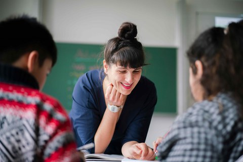 Eine Lehrerin stütze sich auf den Tisch vor zwei Schülern.