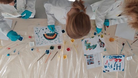 Drei Kinder von oben in weißen Kitteln und blauen Handschuhen, die mit Wasserfarben und Pinsel bunte Bilder malen.