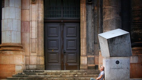 Massives Portal, rechts davor zwei Betonblöcke, daran sitzt ein Mann