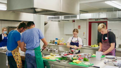 Jugendliche stehen in einer Großküche um einen Tisch und bereiten eine Mahlzeit vor.