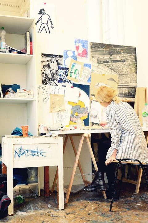 Eine Frau sitzt zeichnend an einem Schreibtisch. Um sie herum stehen und hängen Skizzen und Bilder.