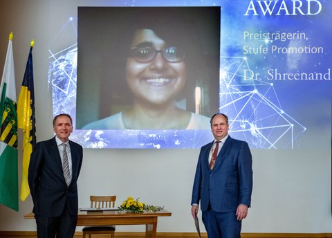 Fotoaufnahme zweier Männer. Im Hintergrund ist eine Projektion einer Videoschalte abgebildet. Sie zeigt das Gesicht einer Frau, die einen Preis der Dresden Excellence Awards gewonnen hat.