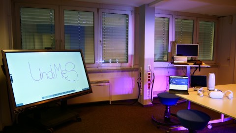 Blick in ein violett ausgeleuchtetes Büro, links ein Monitor mit der Schrift "Undimes" schwarz auf weiß