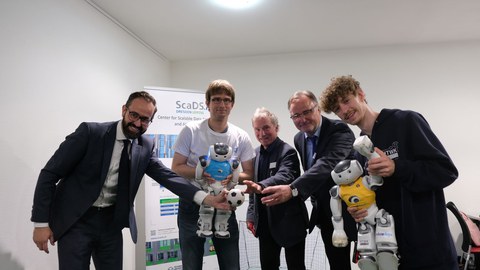 Die fünf genannten Herren stehen nebeneinander, etwas vorgebeugt, und strecken jeweils eine Hand aus.  Hr. Gemkow hält einen kleinen Fußball in der Hand,  Hr. Jagla und Hr. Loos halten jeweils einen kleinen Roboter.