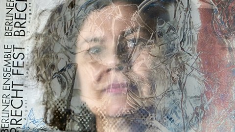 Portrait of a woman behind a structured glass pane, on the left from bottom to top there is lettering reading "Berliner Ensemble, Brecht Fest". 