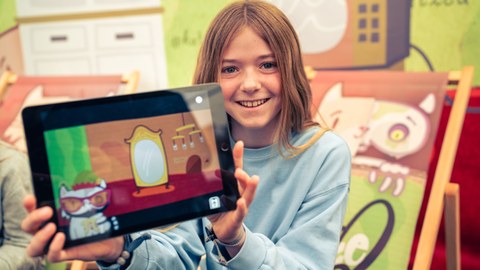 Ein Mädchen zeigt ein Tablet, in dem die App Katze Q zu sehen ist.