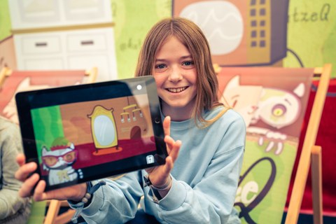 Ein Mädchen zeigt ein Tablet, in dem die App Katze Q zu sehen ist.