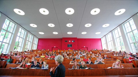 Blick von vorn in einen belegten Hörsaal, im Vordergrund eine Professorin.
