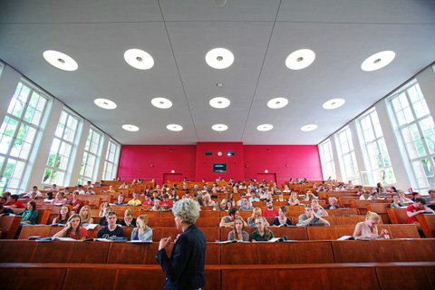 Blick von vorn in einen belegten Hörsaal, im Vordergrund eine Professorin.