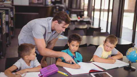 Drei junge Schüler sitzen in einer Reiche am Tisch, ein junger Mann beugt sich zu ihnen und erklärt.