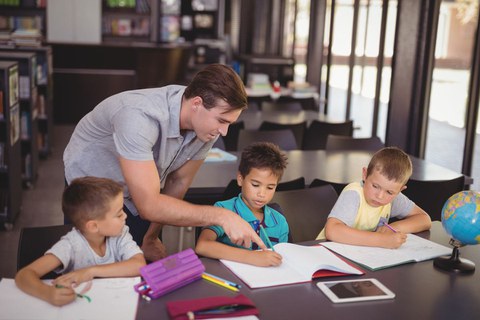 Drei junge Schüler sitzen in einer Reiche am Tisch, ein junger Mann beugt sich zu ihnen und erklärt.