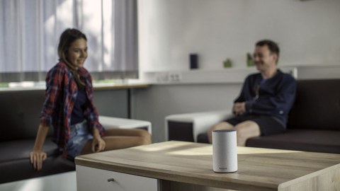 Auf einem Tisch steht ein Lautsprecher, im Hintergrund sitzen eine junge Frau und ein junger Mann auf einem Sofa.