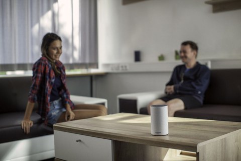 Auf einem Tisch steht ein Lautsprecher, im Hintergrund sitzen eine junge Frau und ein junger Mann auf einem Sofa.