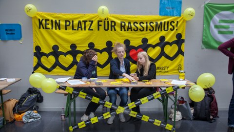 Drei junge Frauen sitzen vor einem gelben Plakat mit der Aufschrift: Kein Platz für Rassismus