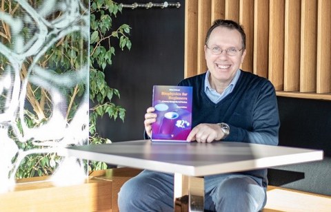 Prof. Schießel sitzt an einem Tisch und hält sein Buch in die Kamera.