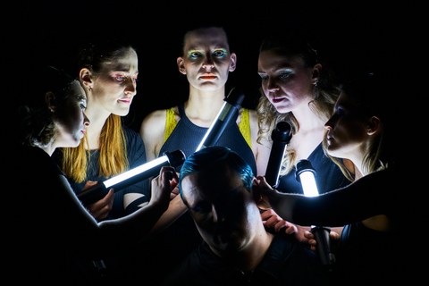 Sechs Personen vor dunklem Hintergrund, fünf der Gesichter sind mit Stablampen erleuchtet. Der Kopf in der Mitte ist im Dunkeln.