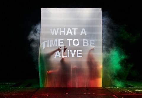 Menschen stehen hinter einem beleuchteten Vorhang, darauf der Text "What a time to be alive".