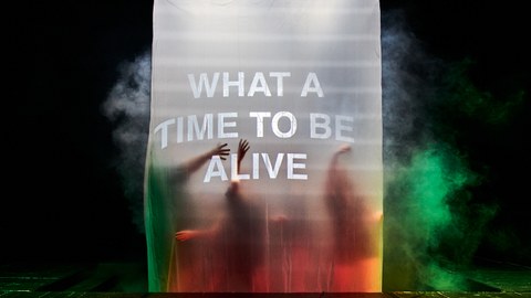 Menschen stehen hinter einem beleuchteten Vorhang, darauf der Text "What a time to be alive".