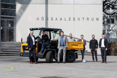 Vor dem Hörsaalzentrum steht ein gelber Dumper. Darum herum gruppiert eine Gruppe Männer.