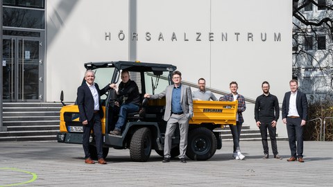 Vor dem Hörsaalzentrum steht ein gelber Dumper. Darum herum gruppiert eine Gruppe Männer.