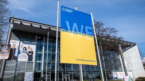 Vor dem Hörsaalzentrum steht ein Banner (oben blau, unten gelb) mit der Aufschrift: We stand with Ukraine