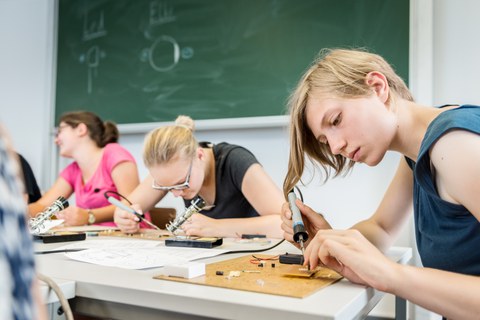 Drei junge Frauen sitzen an Tischen vor einer Tafel und arbeiten mit Lötkolben.