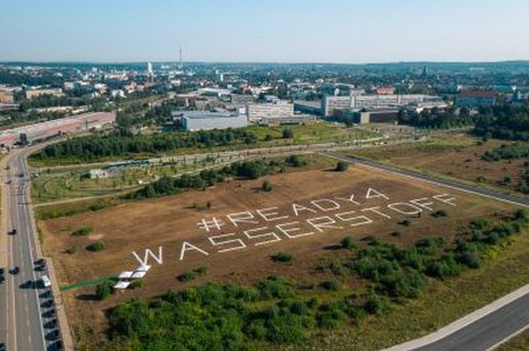 Landschaft, im Hintergrund Industrie. Auf einem Feld die Botschaft "Ready 4 Wasserstoff"