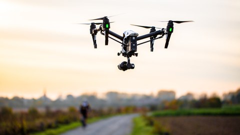 Eine Drohne fliegt über eine Straße, im Hintergrund eine Stadt.