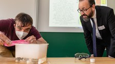 Fotoaufnahme von zwei Männern deren Blicke auf einen großen Würfel aus Trockeneis gerichtet sind, der sich auf einem Tisch befindet.
