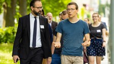 Fotoaufnahme von mehreren Personen, die sich im Freien fortbewegen. Im Vordergrund sind zwei Männer abgebildet. Im Hintergrund befinden sich weitere Personen.