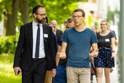Fotoaufnahme von mehreren Personen, die sich im Freien fortbewegen. Im Vordergrund sind zwei Männer abgebildet. Im Hintergrund befinden sich weitere Personen.