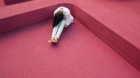 Eine junge Frau sitzt mit hängendem Kopf in einer Ecke auf rotem Teppichboden.