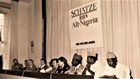 Schwarz-weiß-Foto von Menschen verschiedener Kulturen, die auf einem Podium sitzen. Im Hintergrund ein Banner mit der Aufschrift: Schätze aus Alt-Nigeria, Erbe von 2000 Jahren
