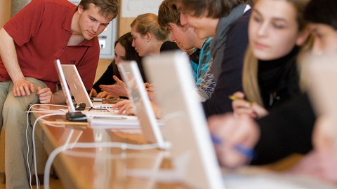 Eine Gruppe von Schülern sitzt vor einer langen Tafel, auf der Monitore stehen.