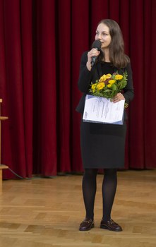 Foto der Preisträgerin Ianina Scheuch, die in ein Mikrofon spricht und eine Urkunde sowie einen Blumenstrauß in den Händen hält.