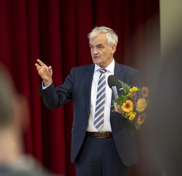 Foto des Preisträgers Prof. Stamm vom ABCD-Centre, der in ein Mikrofon spricht und einen Blumenstrauß in den Händen hält.