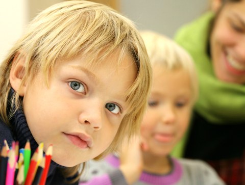Ein blondes Kind schaut von links in die Kamera. Im Vordergrund Buntstifte, im Hintergrund ein weiteres Kind und eine Erwachsene, die sich über dessen Schulter beugt.